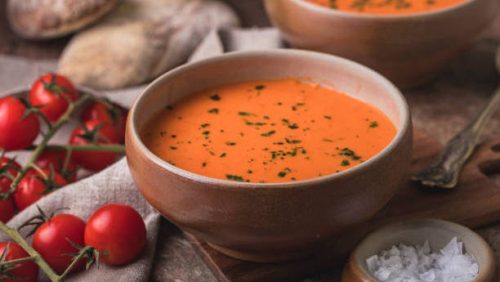 Tomato soup still life with rustic ciabatta bread and fresh tomatoes. The bowl in focus is on a small wooden cutting board. There is a linen cloth next to the bowl, and a small bowl of sea salt.